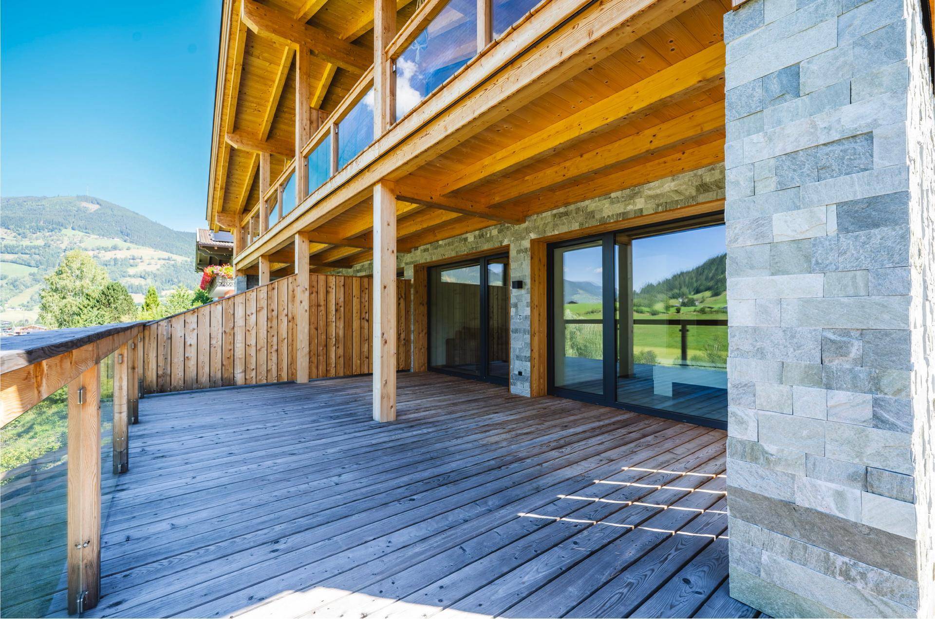 Geräumiger Holzbalkon mit Glasgeländer und Steinwand, bietet einen weiten Blick auf die Berge.