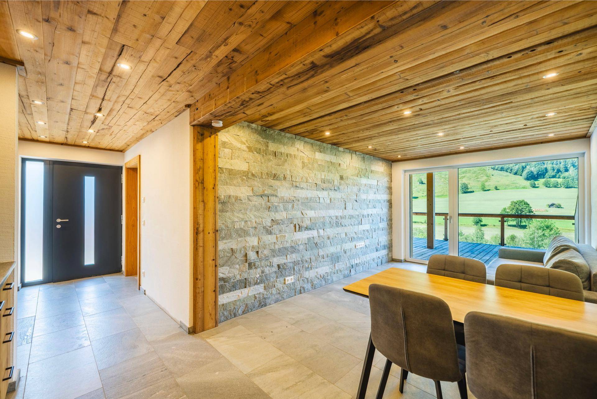 Heller Wohn- und Essbereich mit Holzbalkendecke, Steinwand und Zugang zu einem Balkon mit Bergblick.