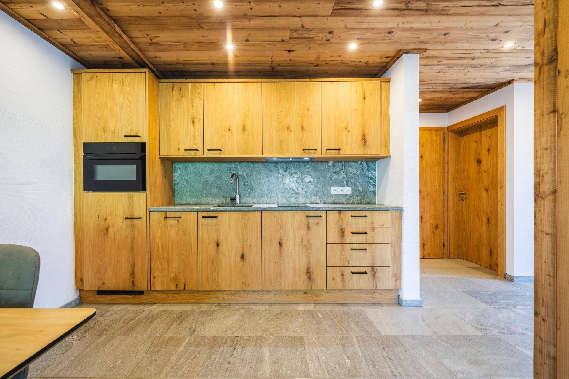 Moderne Holzküche mit Einbaugeräten, Steinrückwand und Fliesenboden in einem offenen Wohnbereich.
