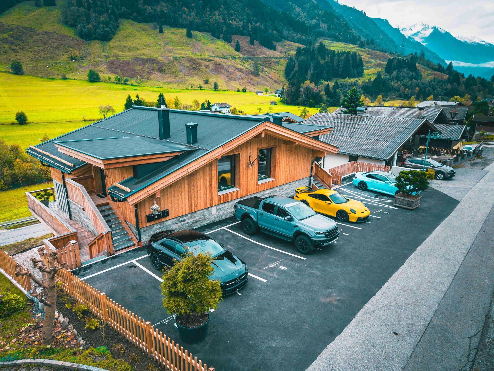 Modernes Holzhaus mit Parkplätzen und Blick auf die umliegende Berglandschaft und grüne Wiesen.