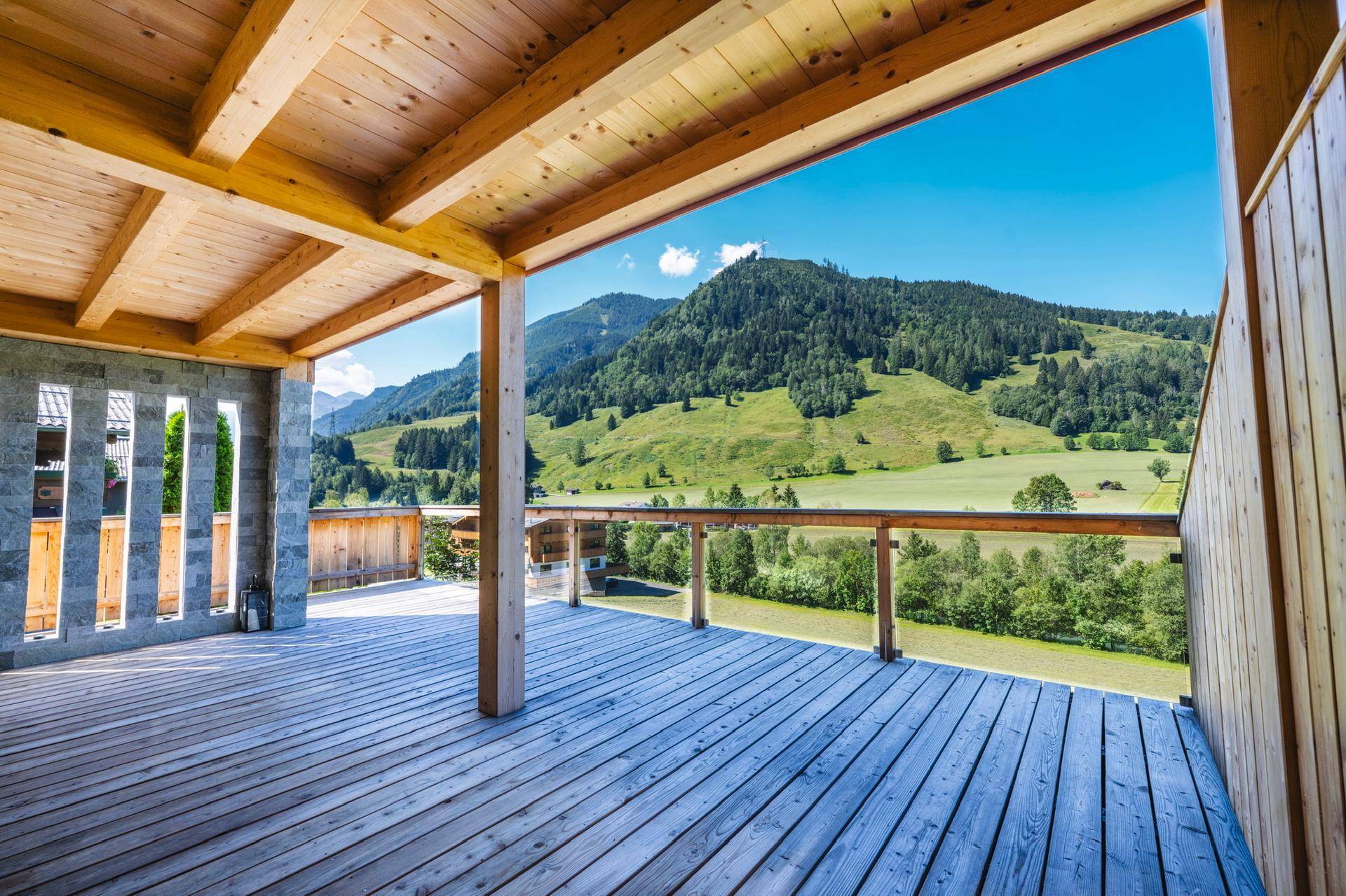 Großer überdachter Holzbalkon mit weitem Blick auf die grüne Berglandschaft und viel Sonne.