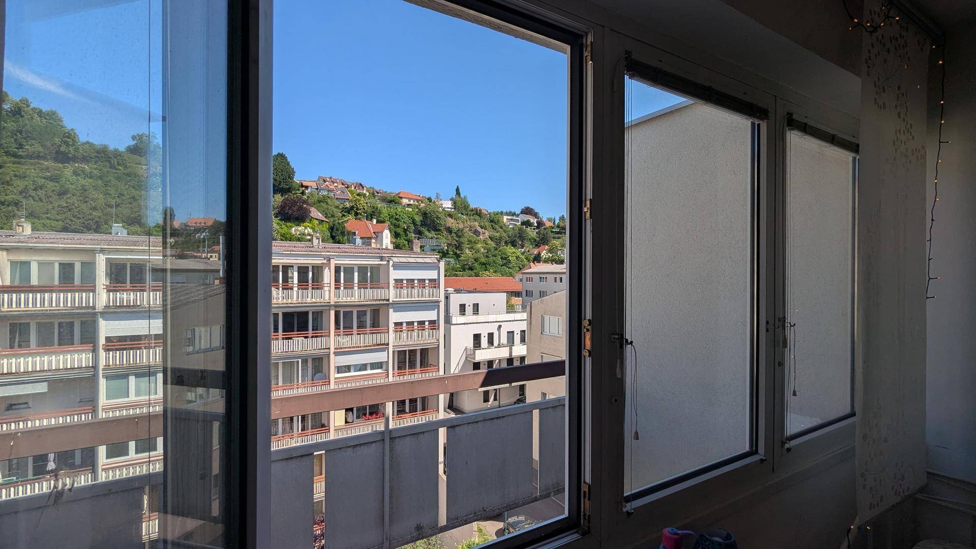 Fensterblick auf die umliegenden Gebäude und einen grünen Hügel unter blauem Himmel.