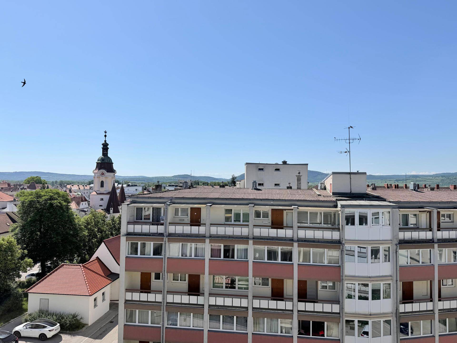 Panoramablick über die Stadt mit Kirchturm und grünen Hügeln im Hintergrund unter blauem Himmel.