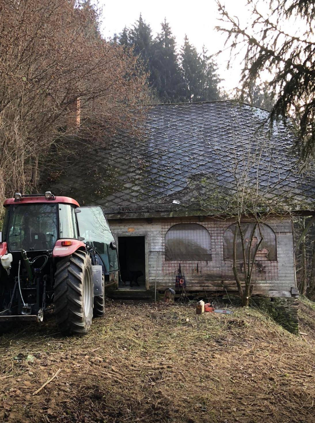 Altes Gebäude mit dunklem Ziegeldach und einem Traktor davor, umgeben von Bäumen.