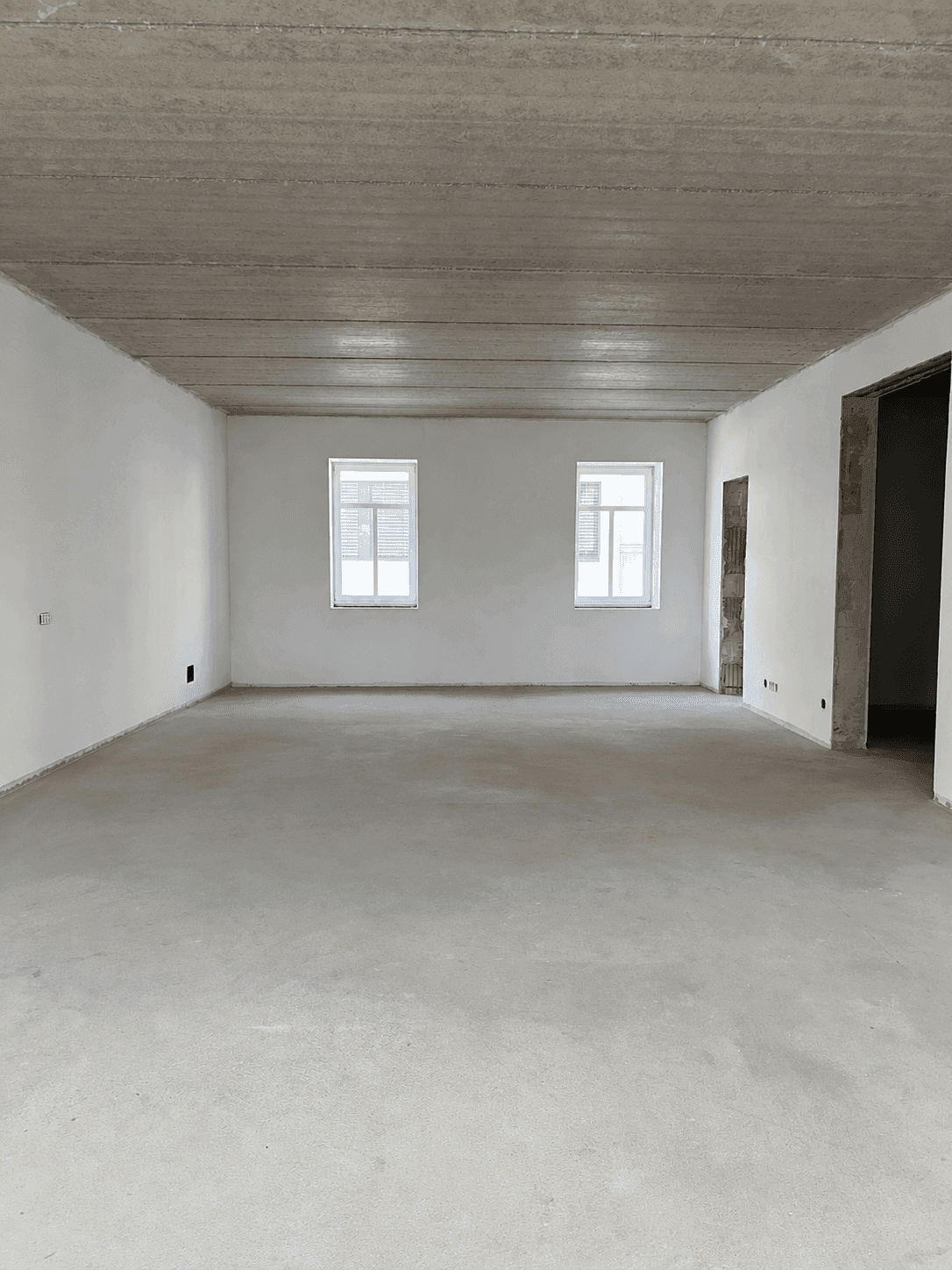Unfertiger Raum mit Betonboden, weißen Wänden und zwei Fenstern, bereit für die Gestaltung.