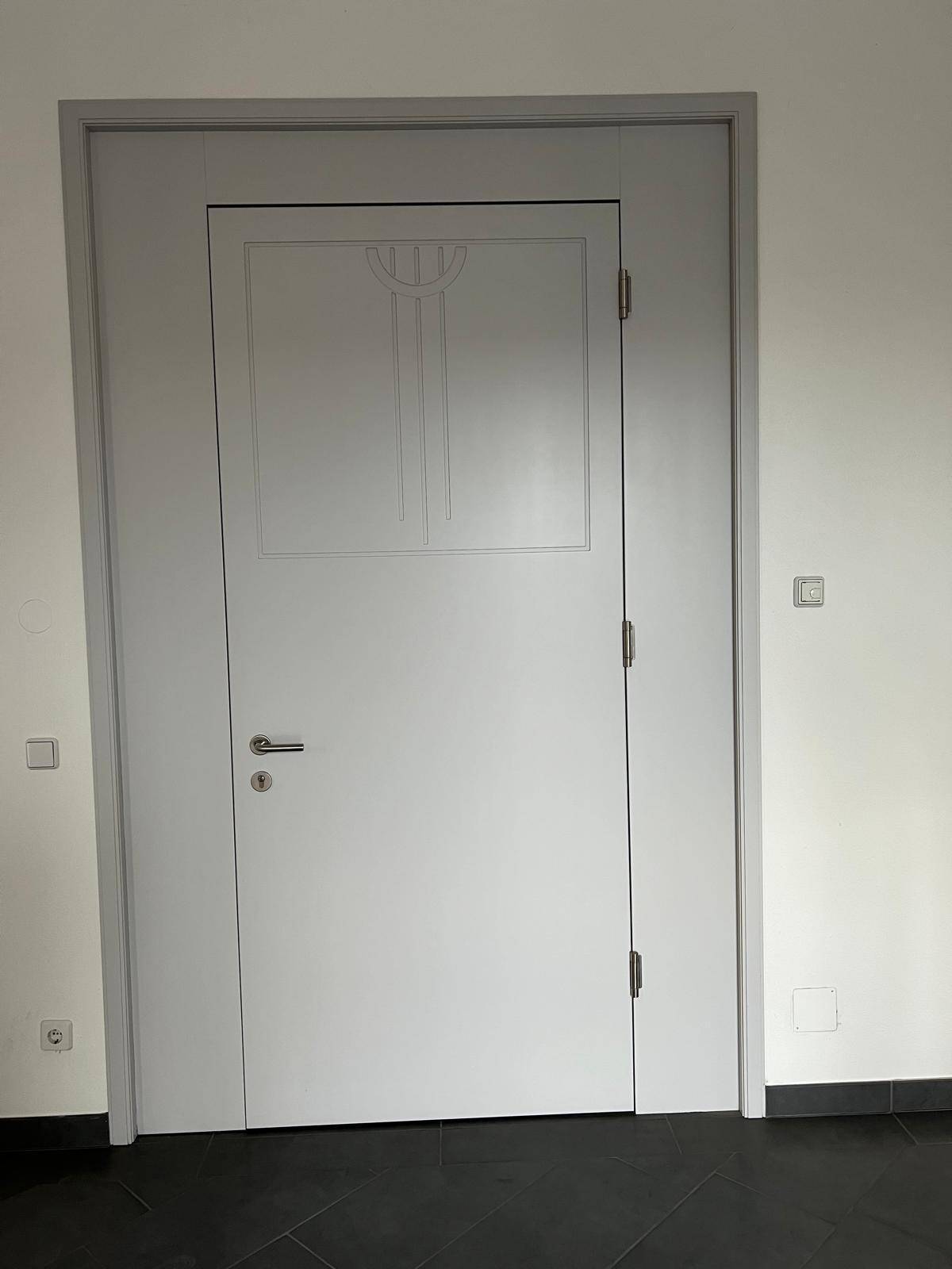 Moderne weiße Tür mit dekorativem Paneel in einem hellen Innenraum mit dunklem Boden.