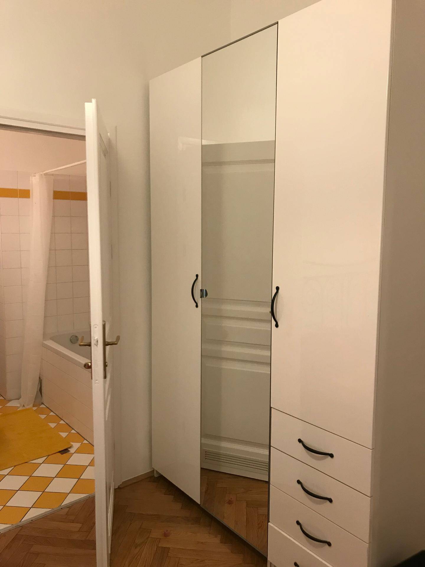 Weißer Kleiderschrank mit Spiegeltüren, dahinter ein Badezimmer mit gelben Fliesen.
