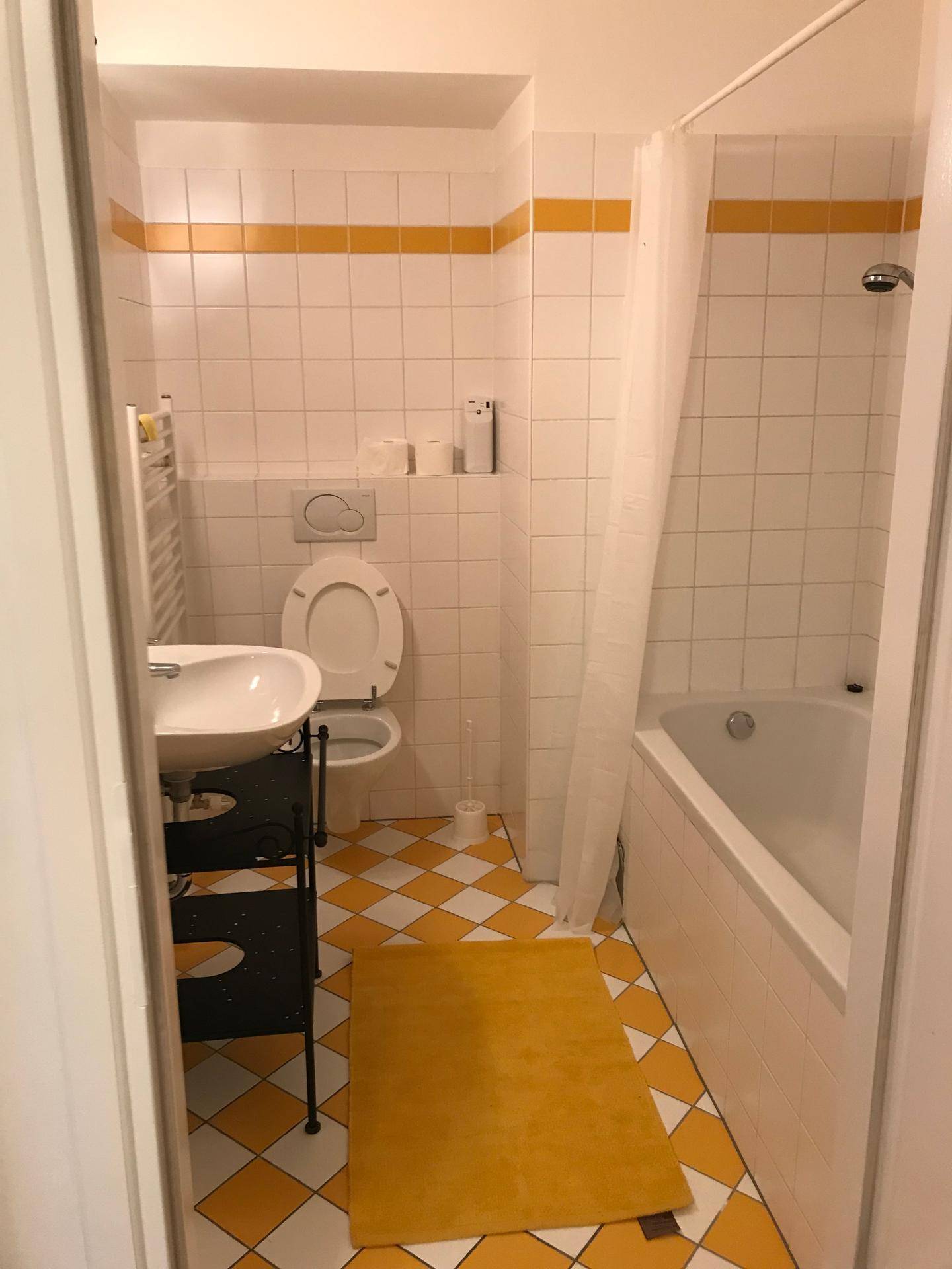 Helles Badezimmer mit Badewanne, WC, Waschbecken und gelb-weiß karierten Bodenfliesen.