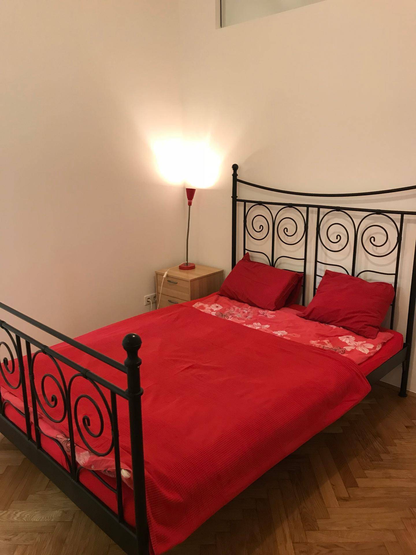 Schlafzimmer mit schmiedeeisernem Bett, roter Bettwäsche und Fischgrätparkett.