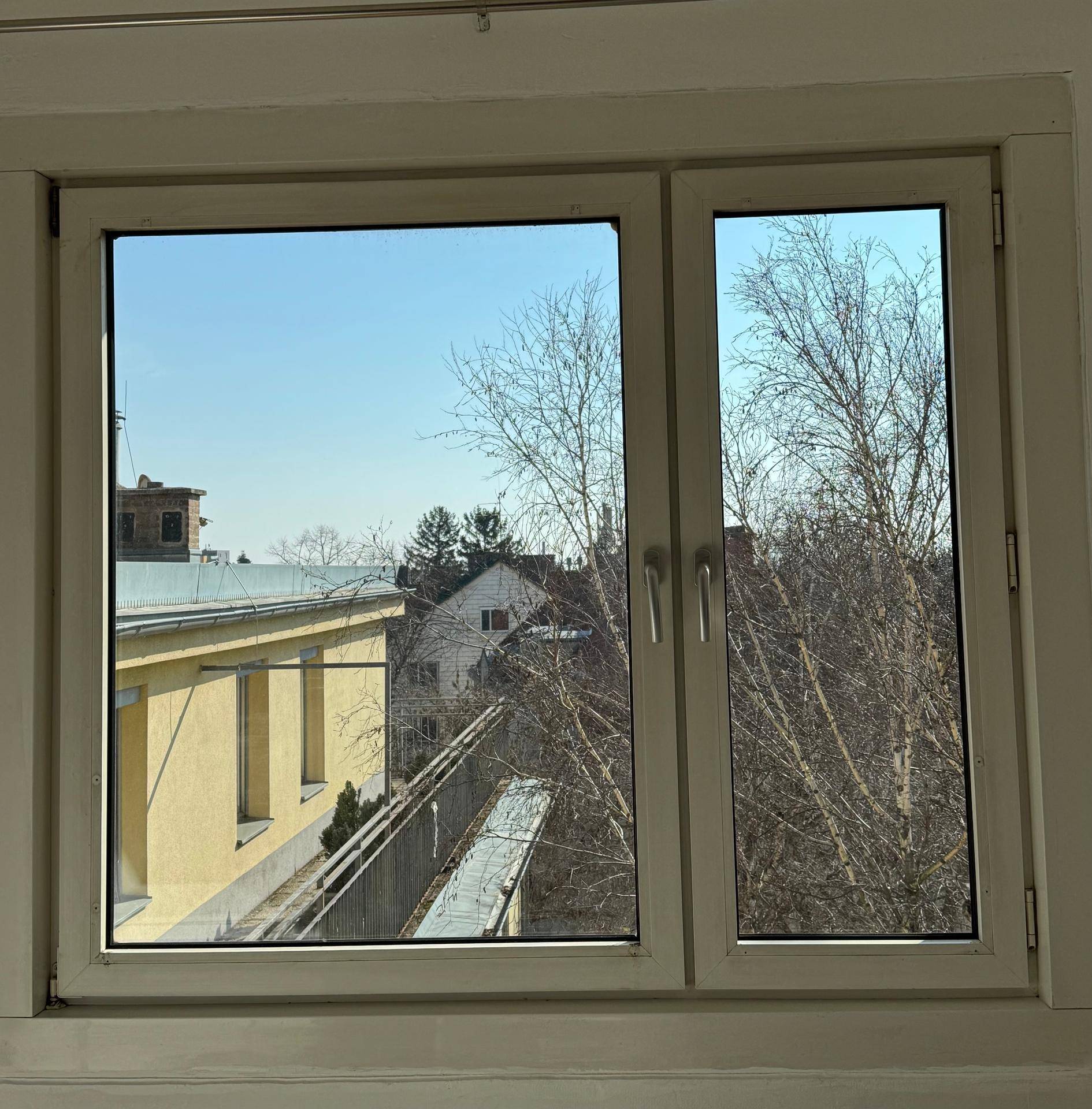 Ausblick aus dem Fenster auf umliegende Gebäude und Bäume unter blauem Himmel.