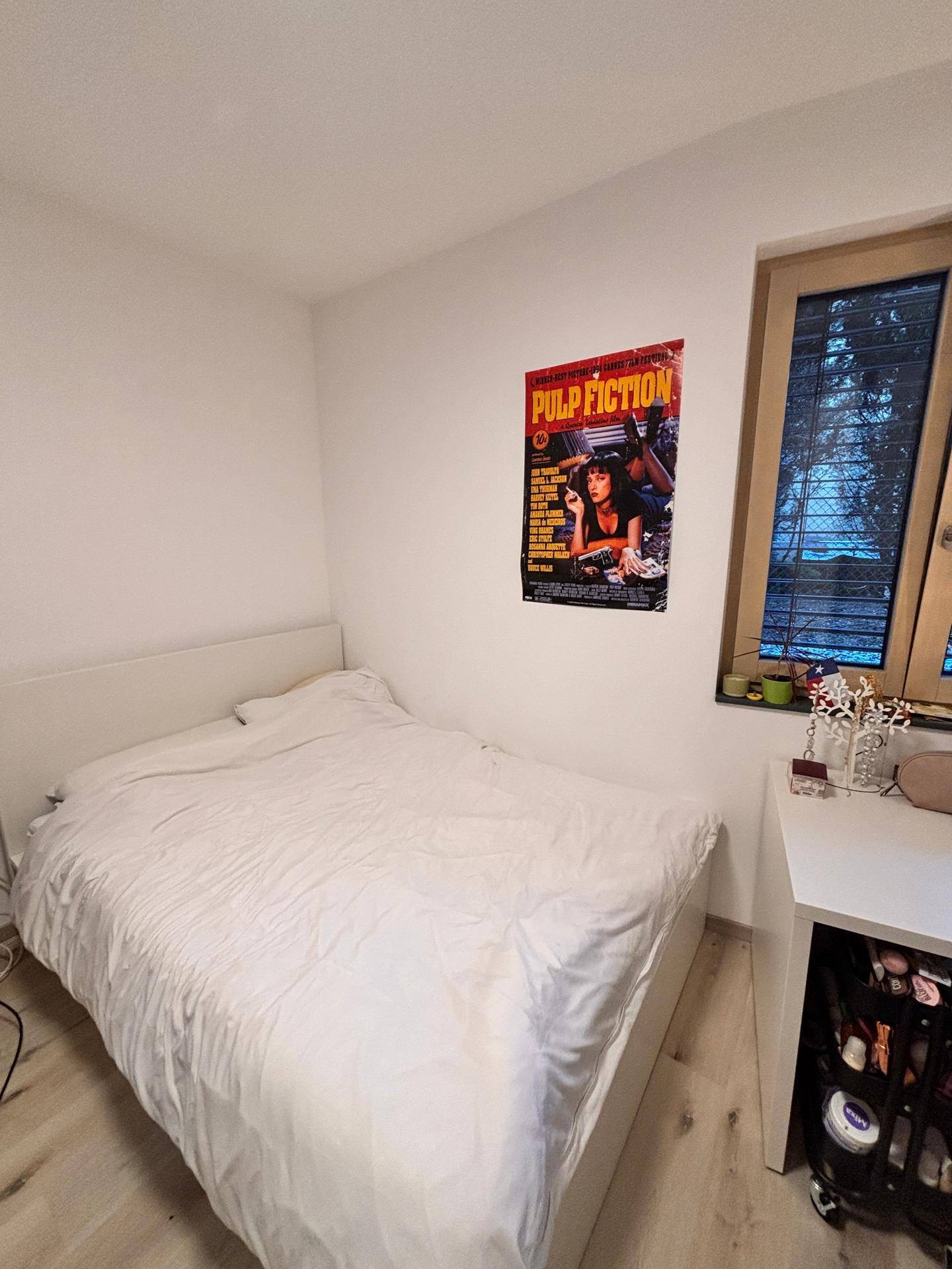 Schlafzimmer mit weißem Bett, Schreibtisch und Fenster mit Jalousien.