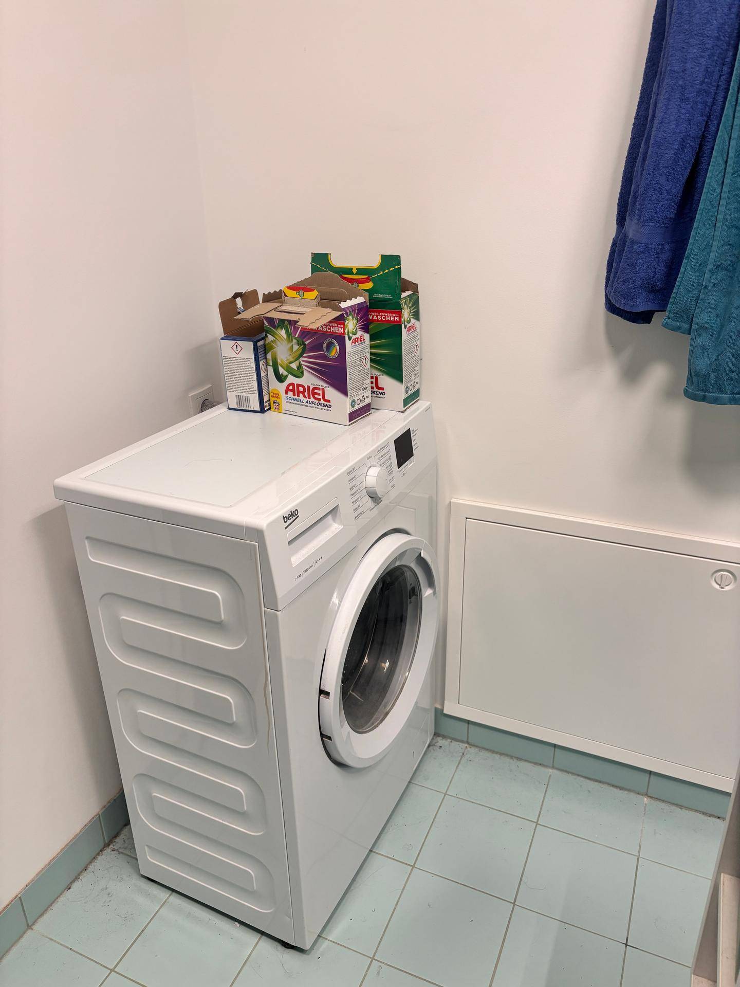 Waschmaschine in einem Bereich mit Fliesenboden und Handtüchern an der Wand.