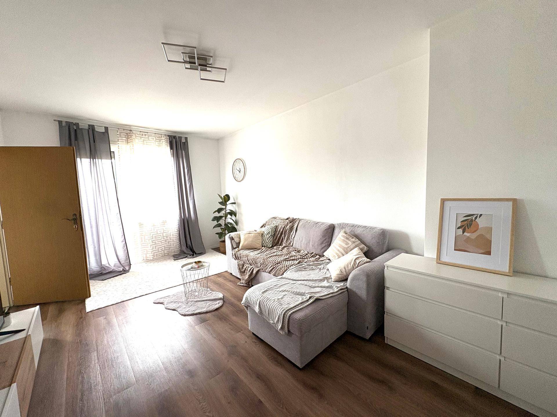 Helles Wohnzimmer mit beigem Sofa, weißer Kommode und Laminatboden, gut beleuchtet durch große Fenster.