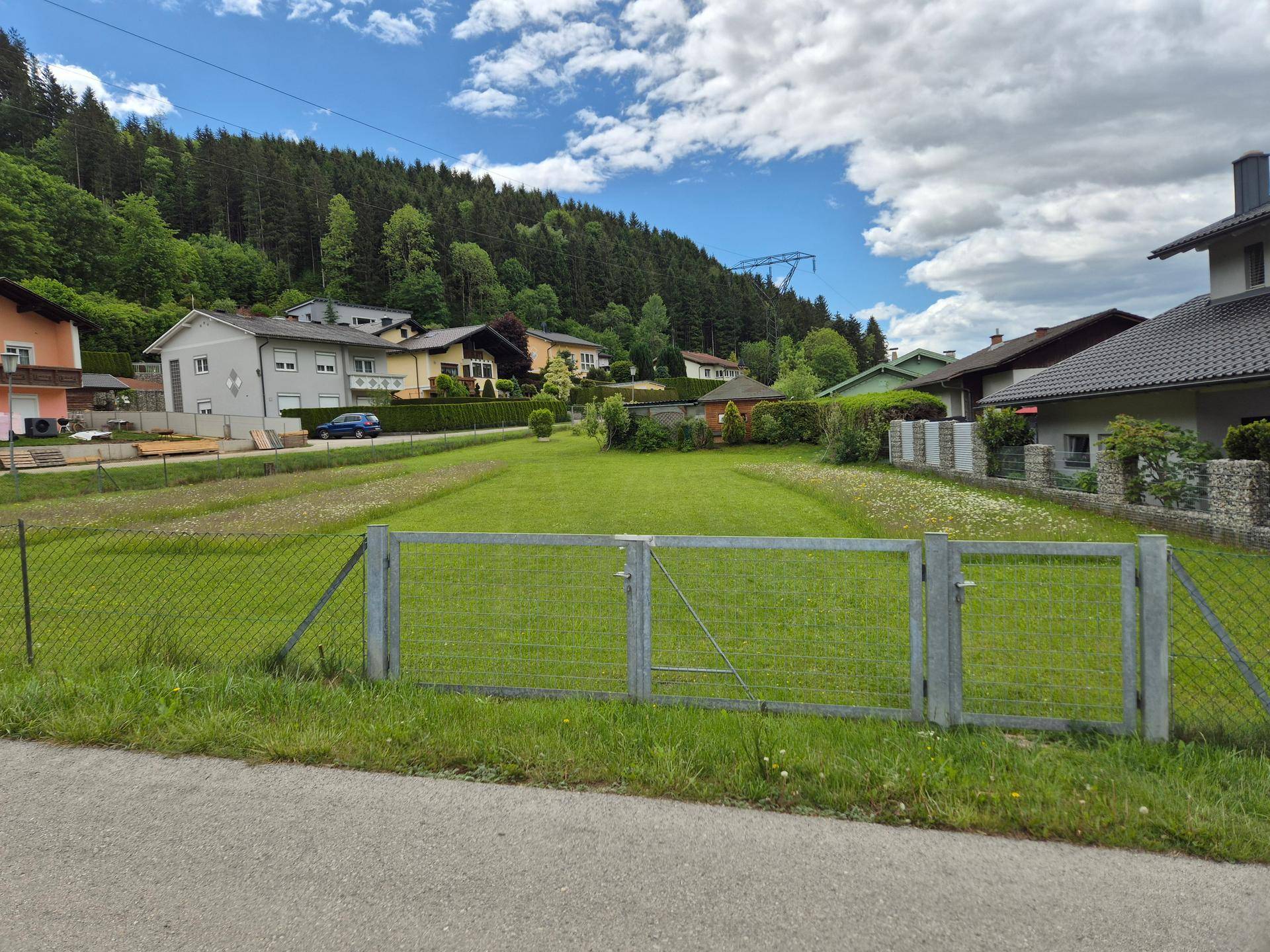 Weitläufiges grünes Grundstück mit Zaun, umgeben von Häusern und bewaldeten Hügeln.