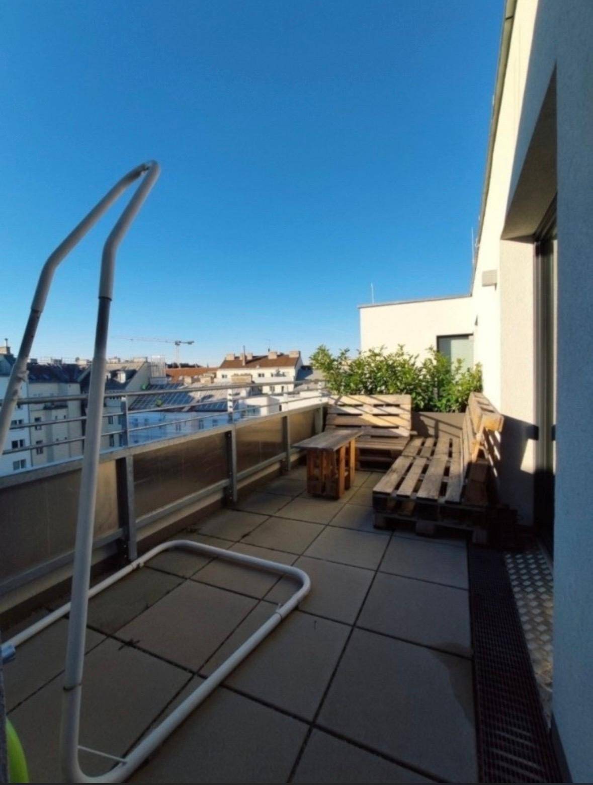 Sonniger Balkon mit Stadtblick und selbstgebauten Palettenmöbeln unter blauem Himmel.