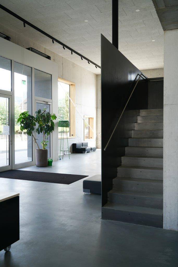 Heller Eingangsbereich mit Glastüren, einer modernen Betontreppe und hohen Decken.