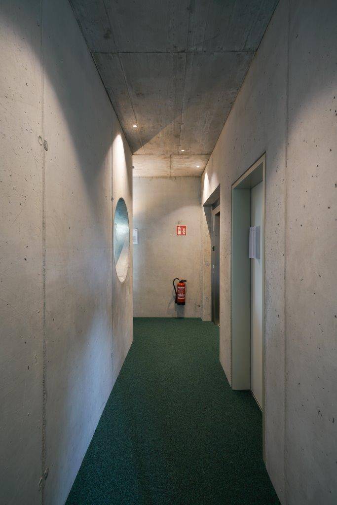 Langer Flur mit Sichtbetonwänden, grünem Teppichboden und einem Aufzug am Ende.
