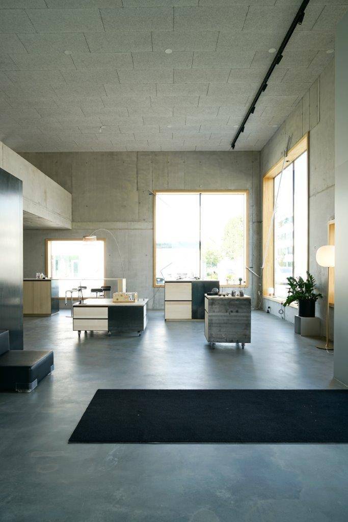 Weitläufiger, heller Raum mit Betonwänden und -decken, großen Fenstern und minimalistischer Einrichtung.