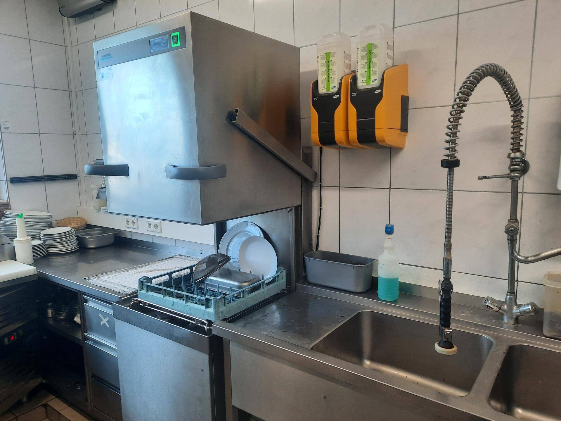 Spülküche mit großem Industriegeschirrspüler, Edelstahlspüle und Wandregalen für Geschirr.