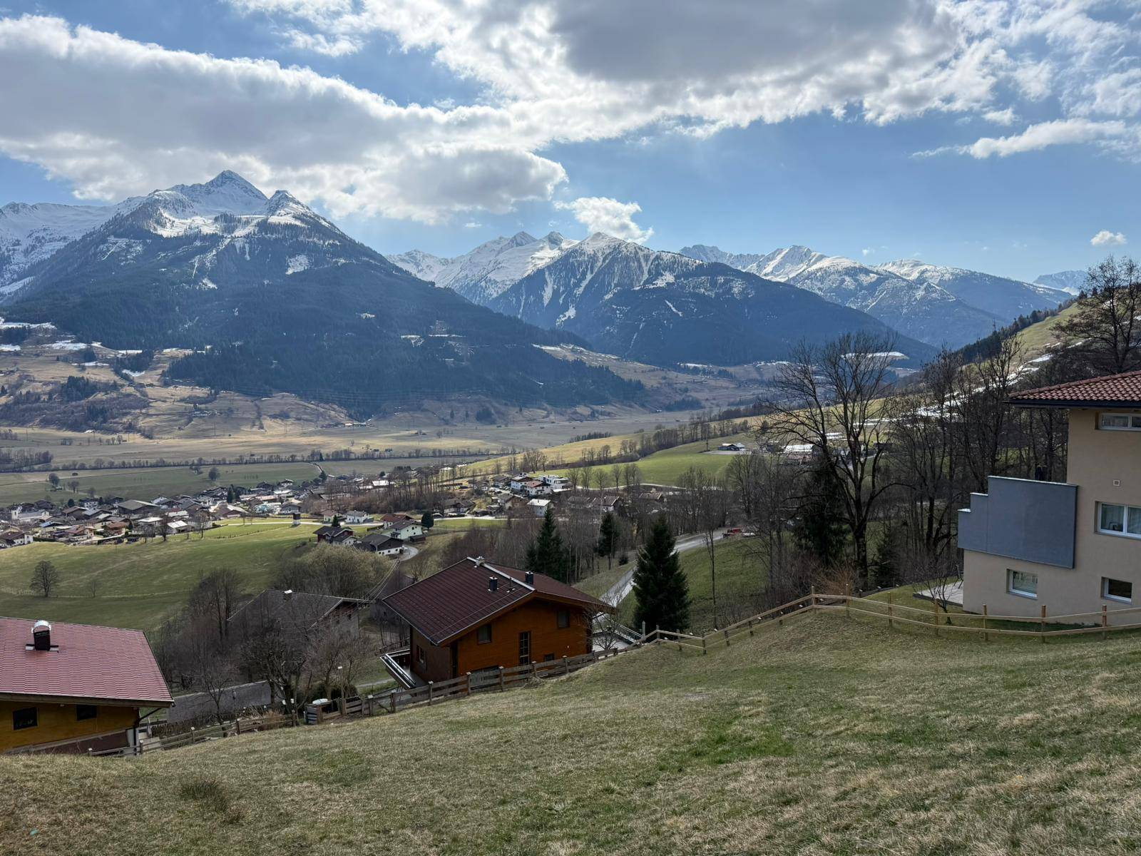 Panoramablick auf ein idyllisches Tal mit Wohnhäusern, grünen Wiesen und schneebedeckten Gipfeln.