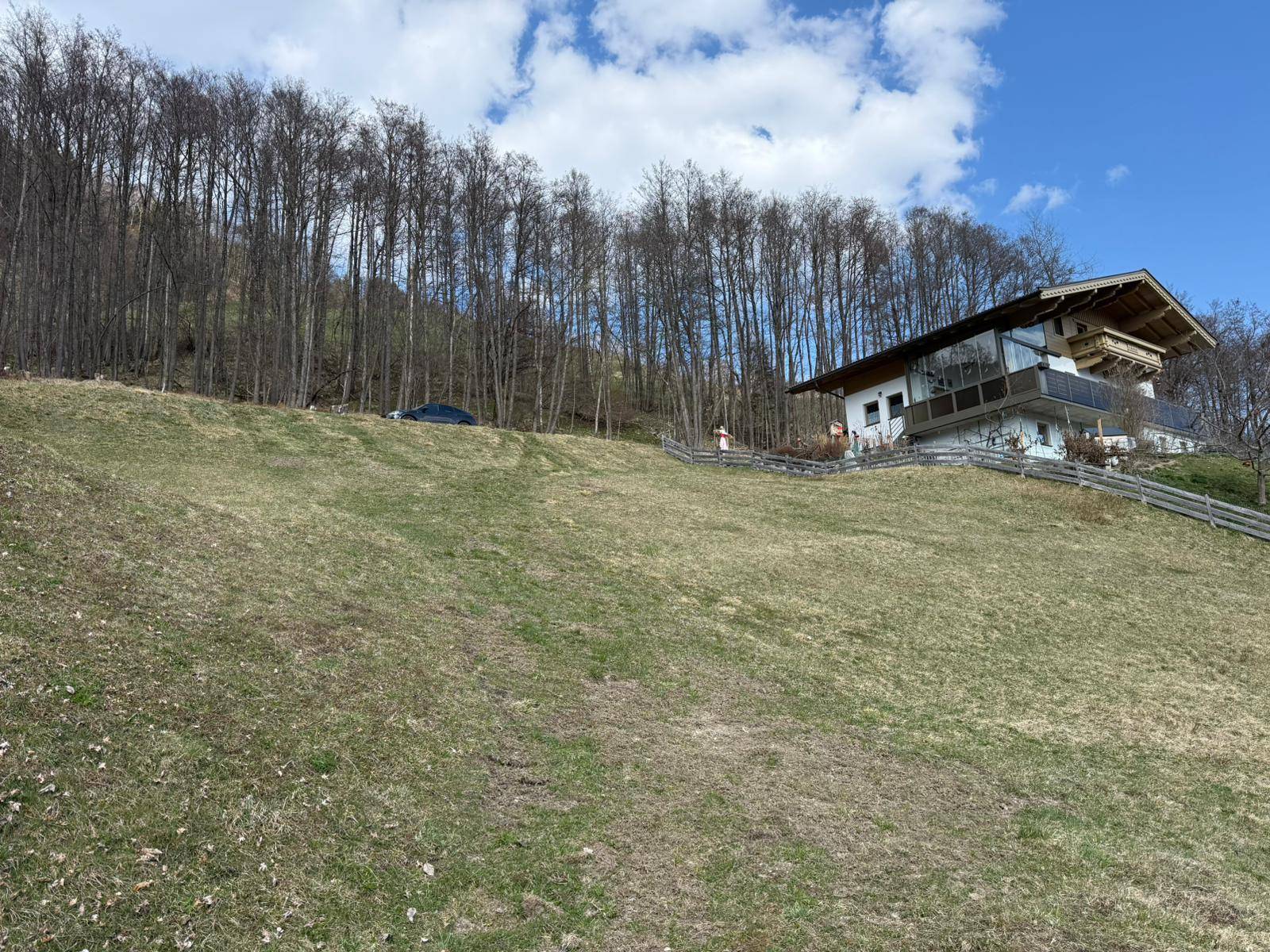 Haus am Hang mit großen Fenstern und Balkonen, umgeben von Bäumen und einer grünen Wiese.