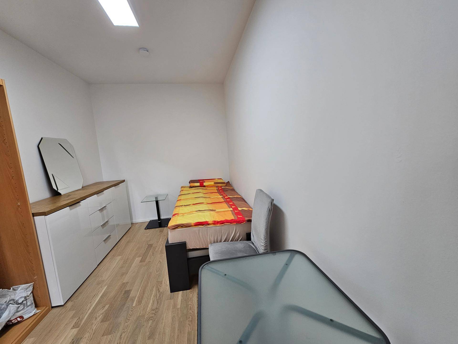 Einfaches Schlafzimmer mit Holzboden, Einzelbett und weißer Kommode mit Spiegel.