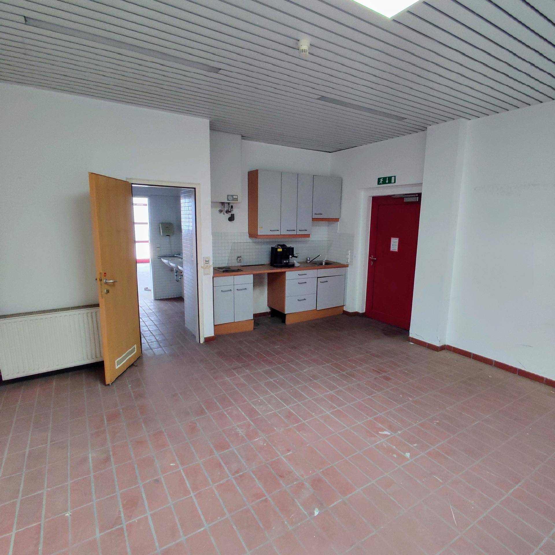 Kleiner Küchenbereich mit grauen Schränken, Arbeitsplatte und roten Bodenfliesen in einem größeren Raum.