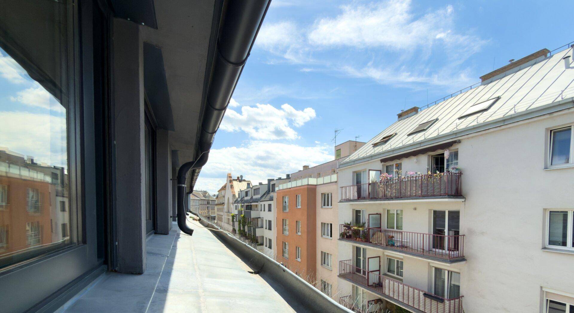 Blick von einem schmalen Balkon auf gegenüberliegende Wohngebäude mit Balkonen und einem weißen Dach.