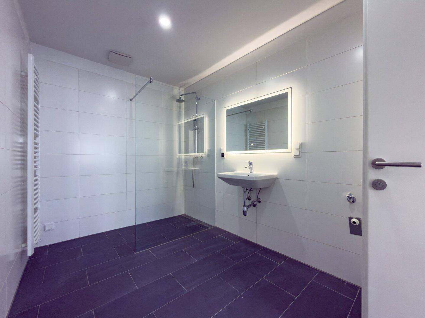 Geräumiges, modernes Badezimmer mit weißen Fliesen, dunklem Boden, begehbarer Dusche und Handtuchheizkörper.