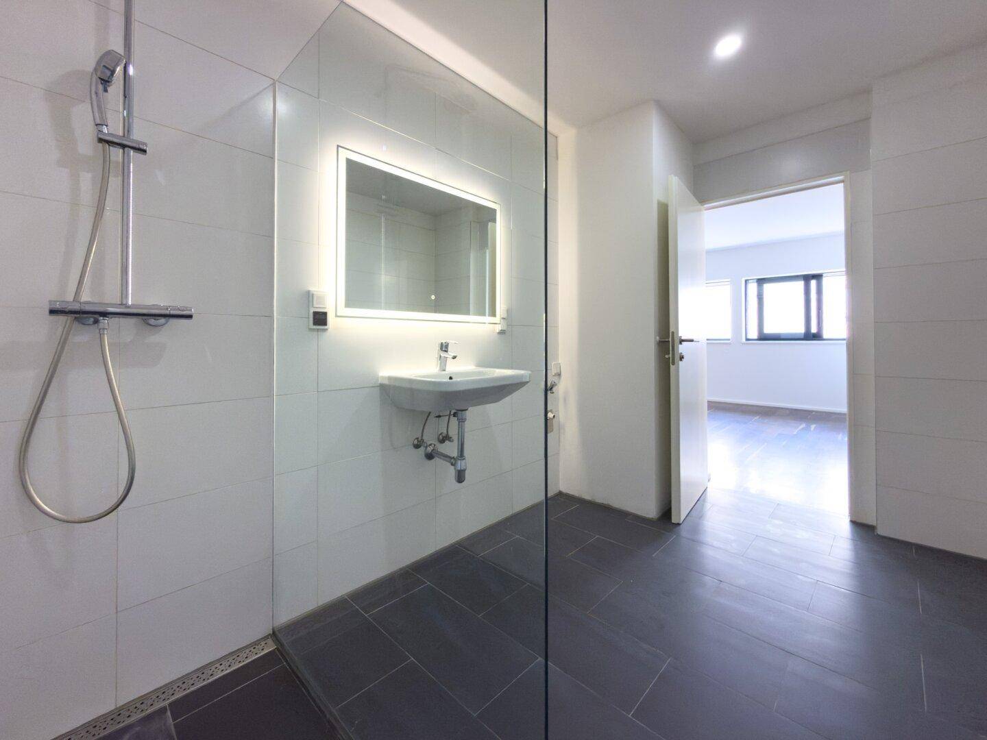 Modernes Badezimmer mit weißen Fliesen, dunklem Boden, wandhängendem Waschbecken und begehbarer Dusche.