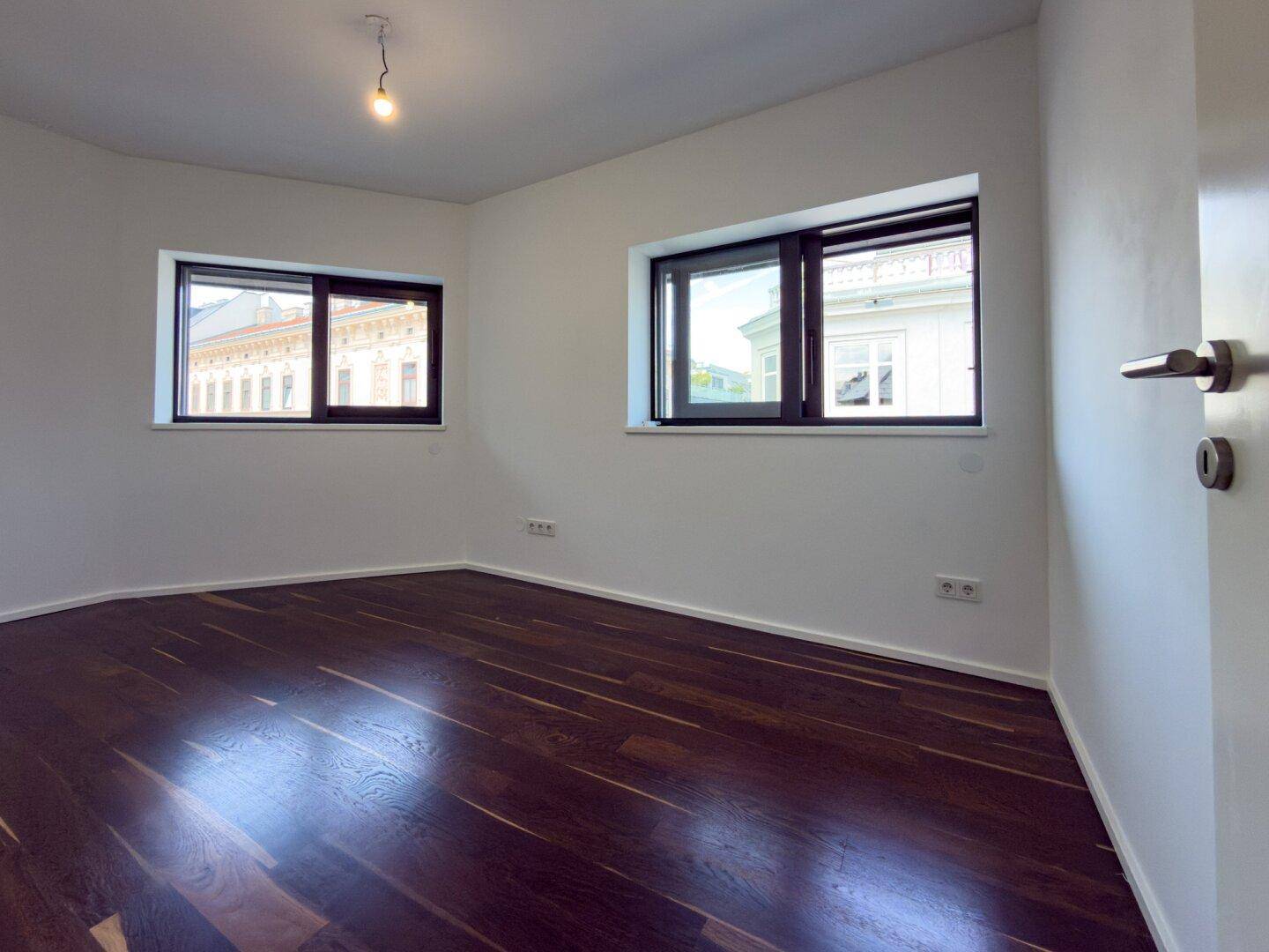 Helles Schlafzimmer mit dunklem Holzboden, weißen Wänden und zwei Fenstern mit Stadtblick.