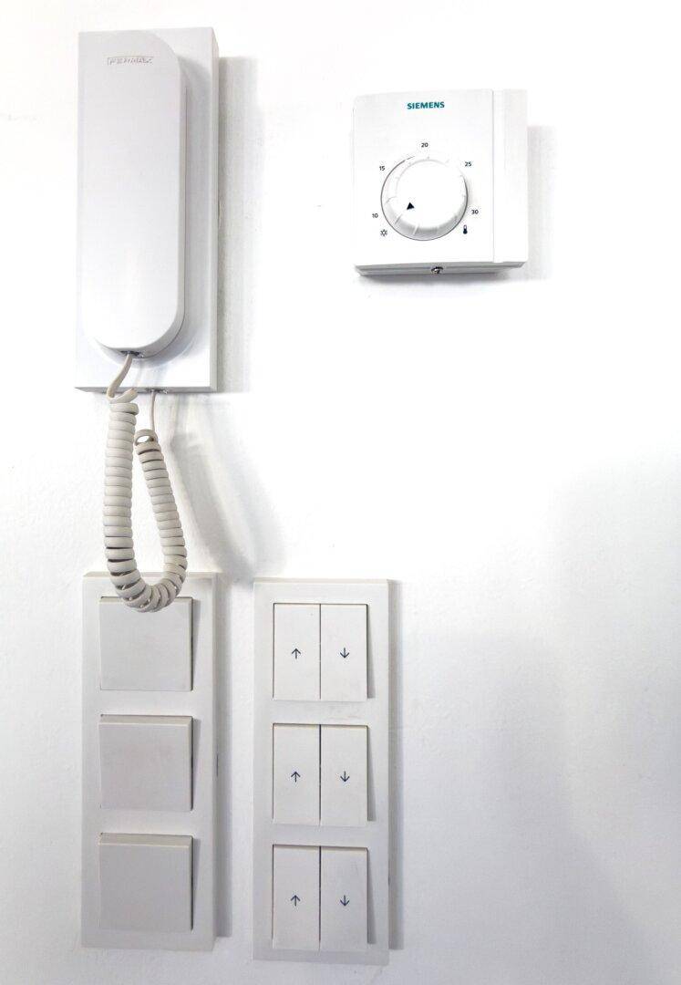 Nahaufnahme einer Gegensprechanlage, eines Siemens-Thermostats und Lichtschaltern an einer weißen Wand.