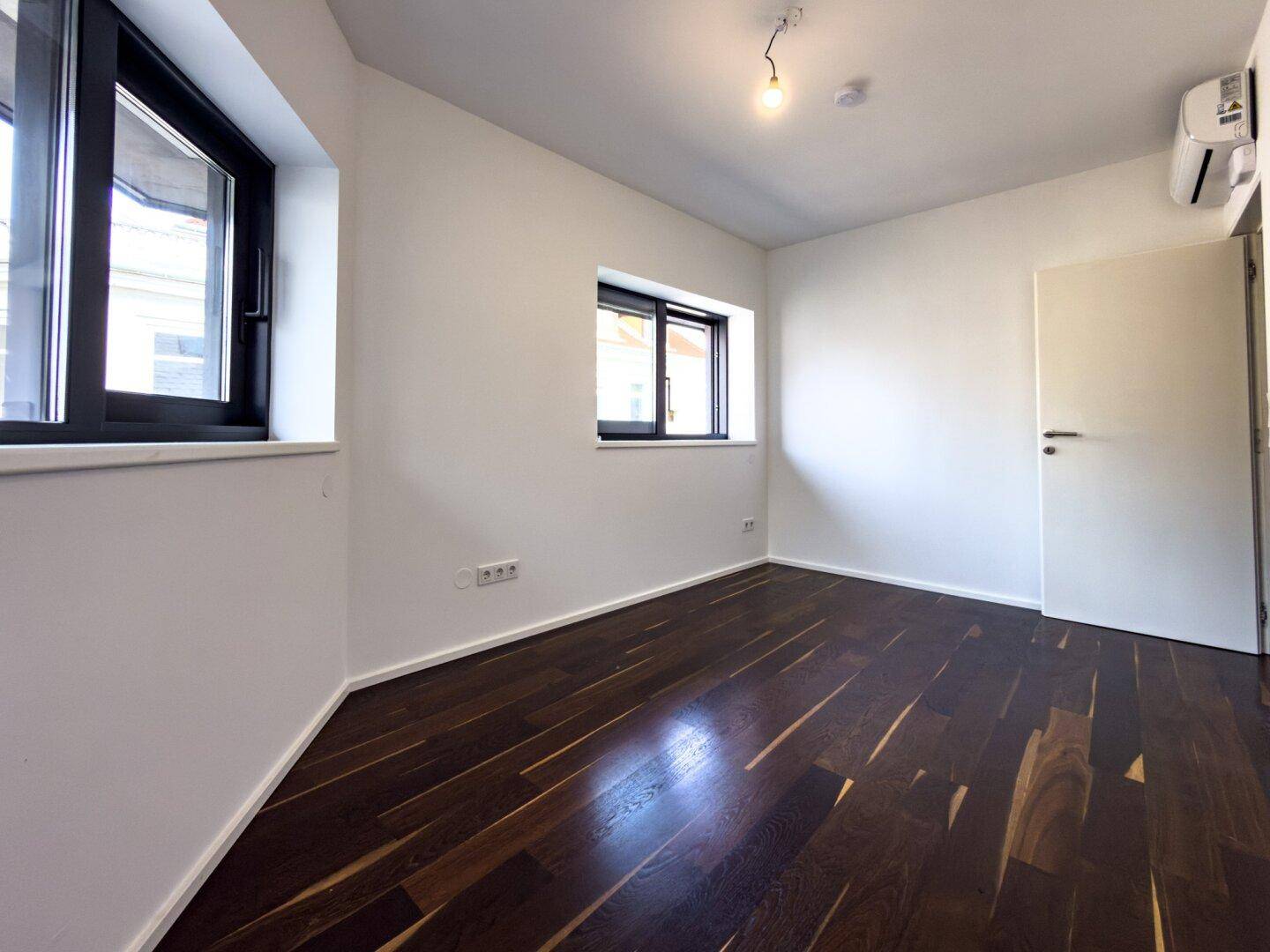 Leeres Schlafzimmer mit dunklem Holzboden, weißen Wänden, zwei Fenstern und Klimaanlage.
