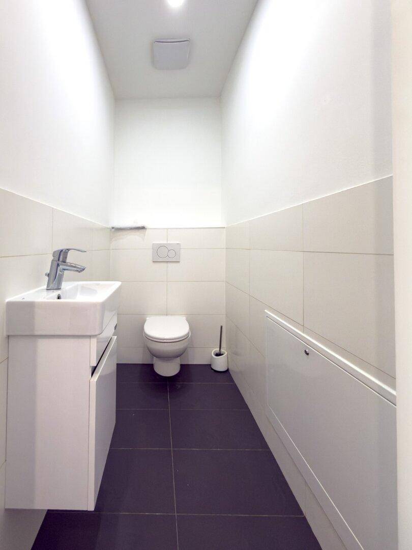 Modernes Gäste-WC mit weißen Fliesen, dunklem Boden, wandhängendem Waschbecken und Toilette.