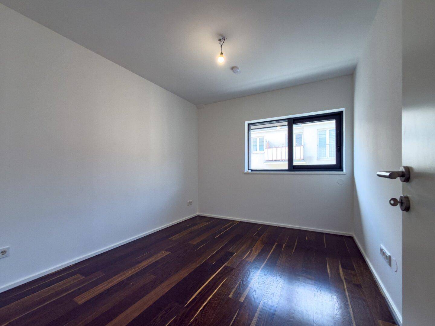Helles Schlafzimmer mit dunklem Holzboden, weißen Wänden und einem Fenster mit Stadtblick.