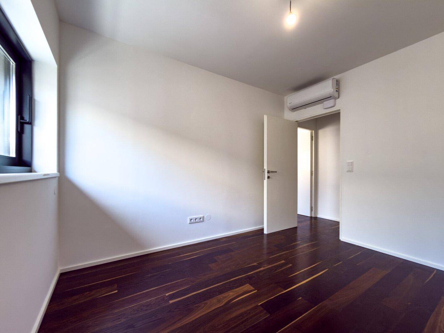 Leeres Schlafzimmer mit dunklem Holzboden, weißen Wänden, einem Fenster und Klimaanlage.