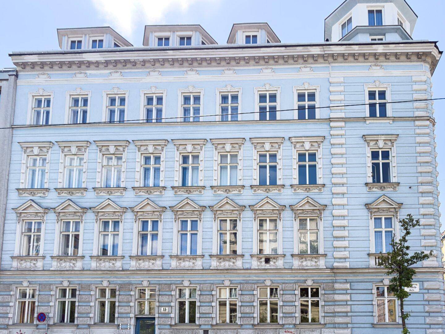 Historische Fassade eines eleganten Gebäudes in Hellblau mit zahlreichen Fenstern und Stuckelementen.