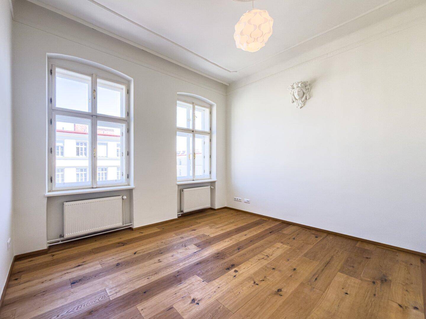 Geräumiges Zimmer mit Parkettboden, hohen Decken und dekorativem Wandelement, bietet viel Licht.