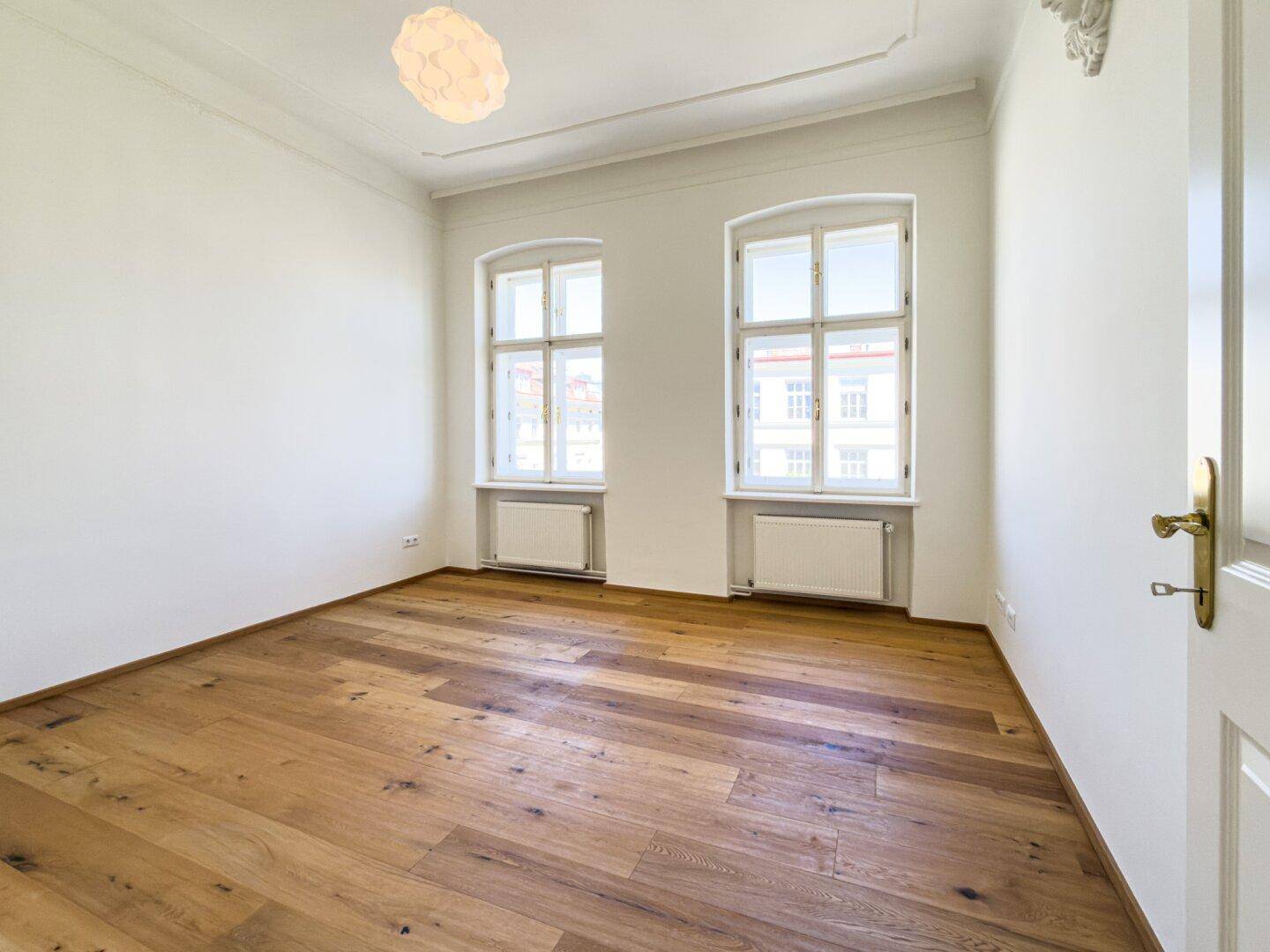 Helles Zimmer mit Holzboden und zwei großen Fenstern, ideal als Schlaf- oder Arbeitszimmer.