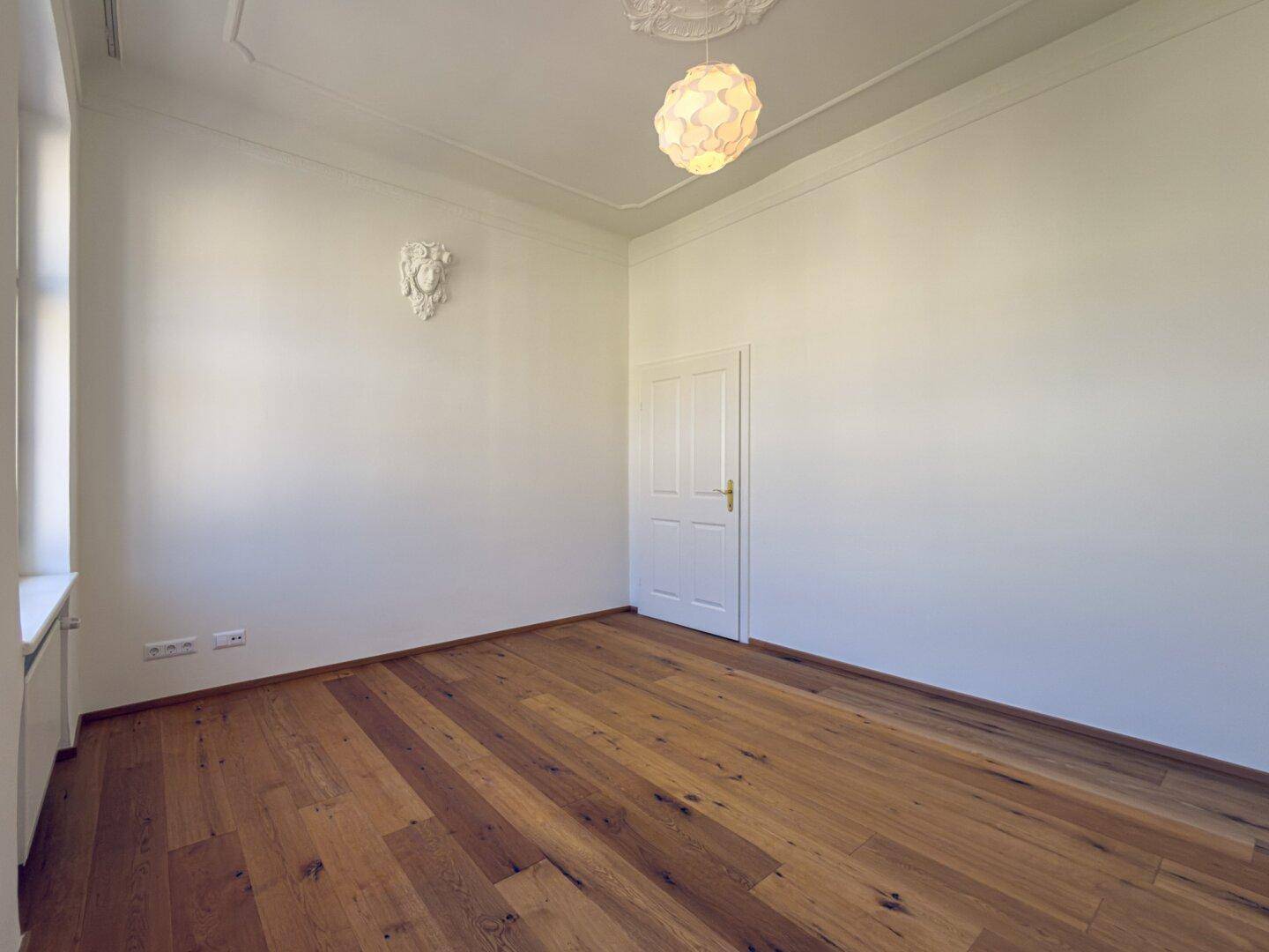 Helles Zimmer mit Holzboden, einer Tür und einem Fenster, bietet eine ruhige Atmosphäre.