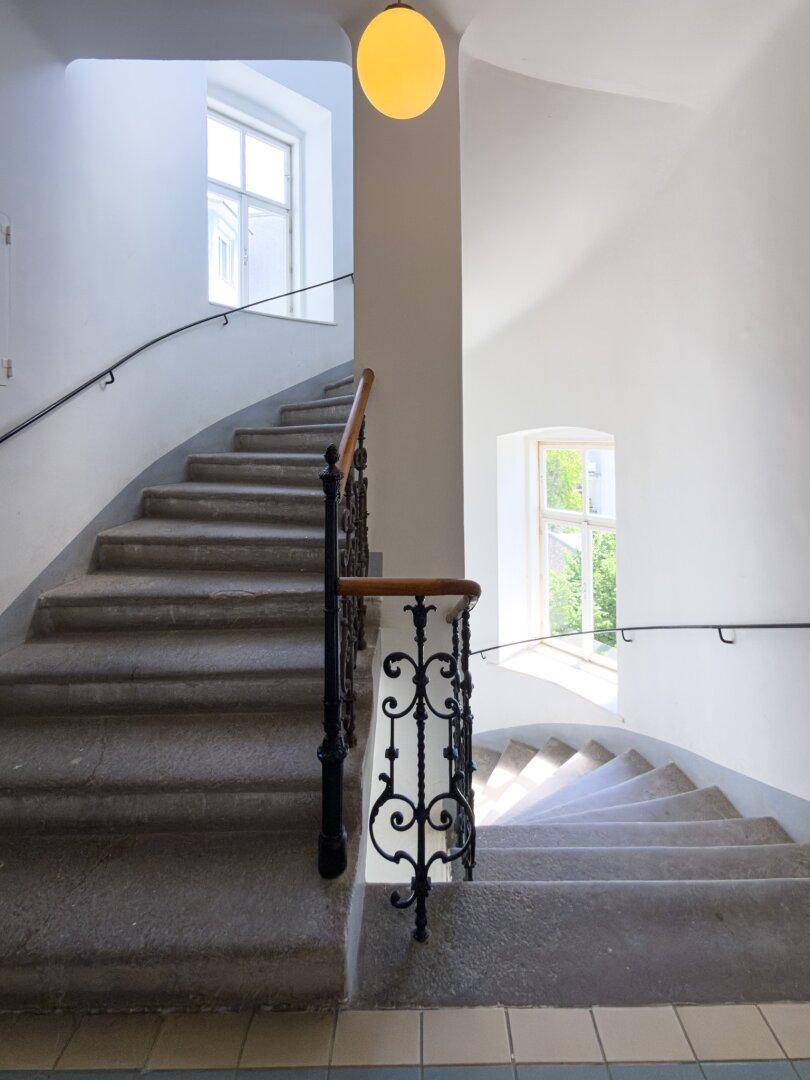 Helles Treppenhaus in einem Altbau mit Steinstufen, schmiedeeisernem Geländer und hohen Decken.