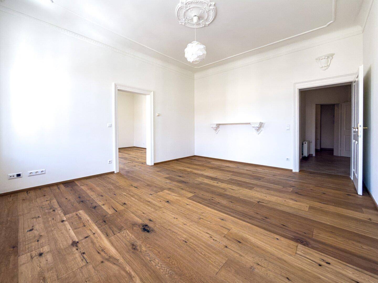 Großes Zimmer mit Parkettboden, hohen Decken und zwei Türöffnungen, ideal für offenes Wohnen.