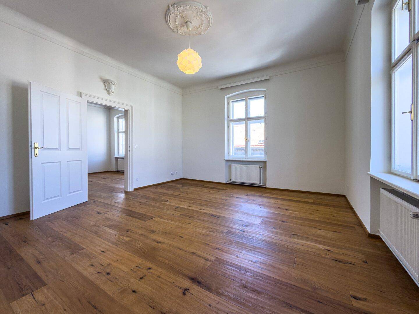 Helles Zimmer mit Parkettboden, hohen Decken und zwei Fenstern, ideal für offenes Wohnen.
