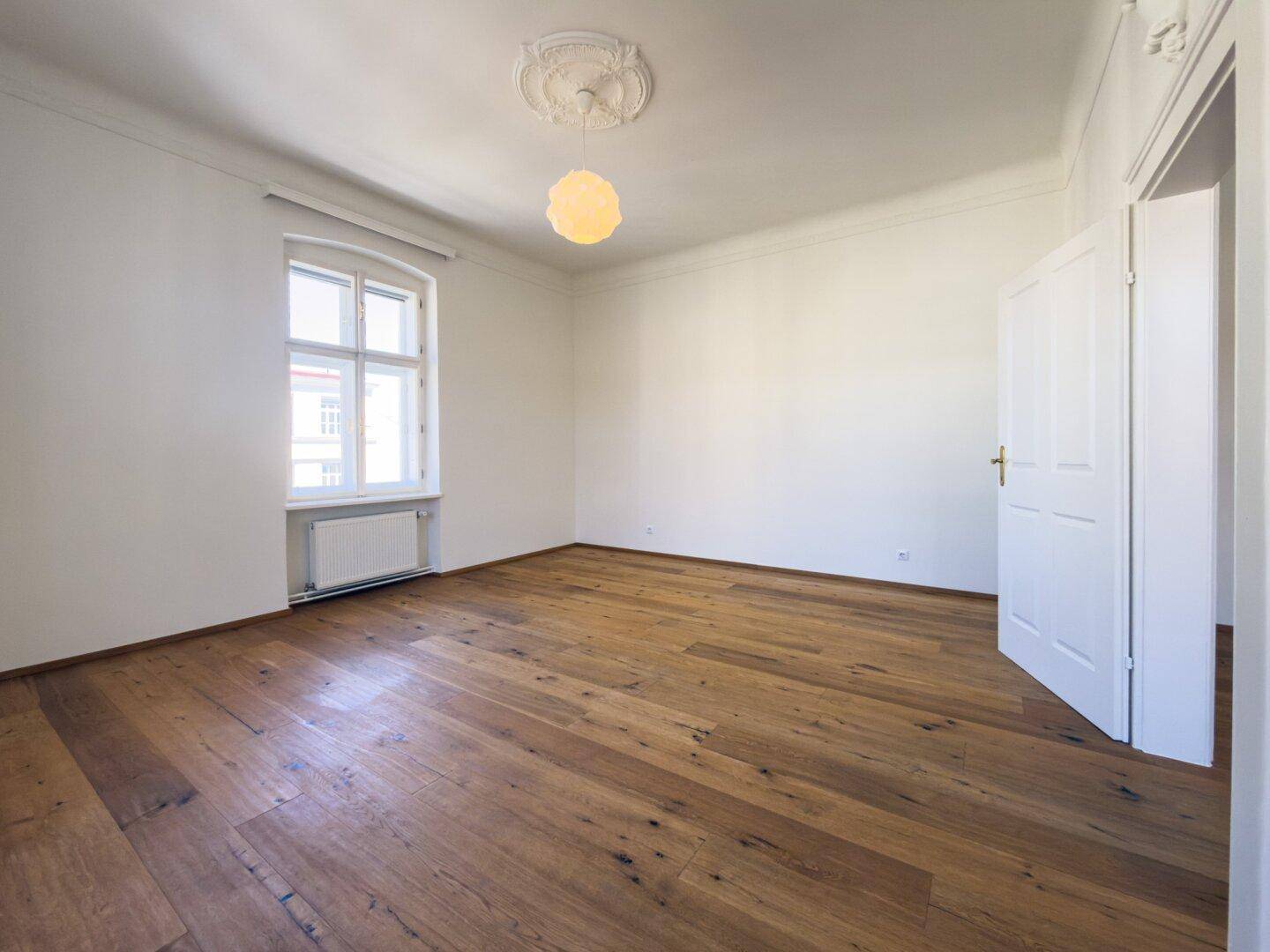 Helles Zimmer mit Holzboden, einem Fenster und einer Tür, bietet eine gemütliche Wohnatmosphäre.