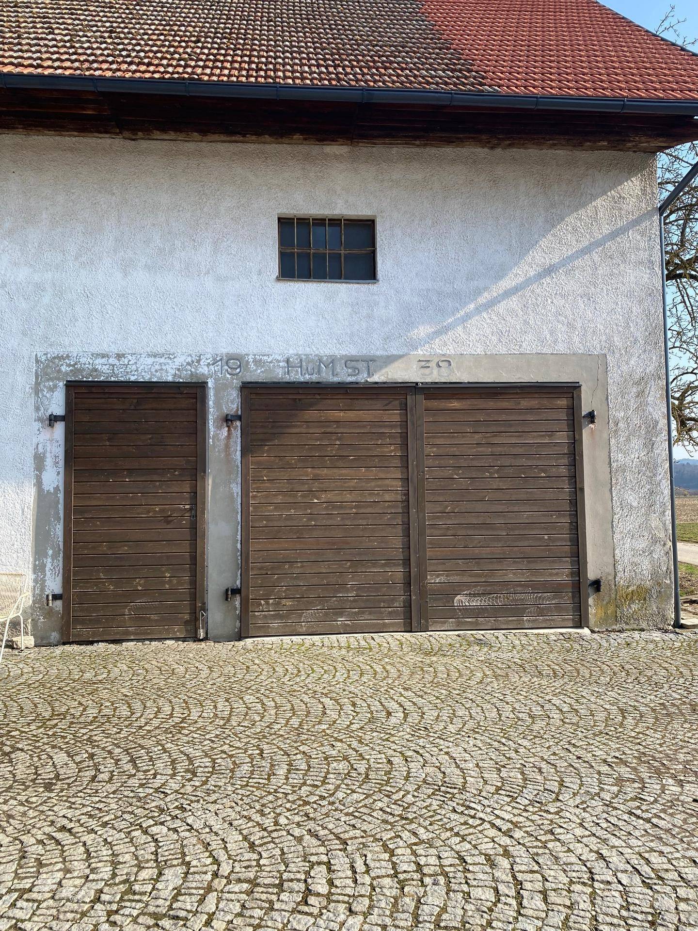 Fassade eines Gebäudes mit kleinen Fenstern und drei großen Holztoren, davor ein gepflasterter Hof.