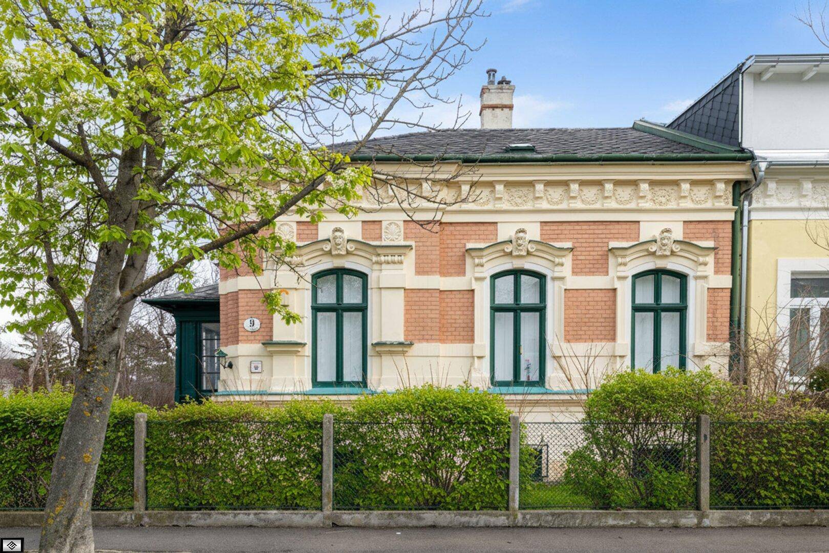Historische Fassade eines Altbaus mit verzierten Fenstern, Ziegelmauerwerk und gepflegter Hecke zur Straße.