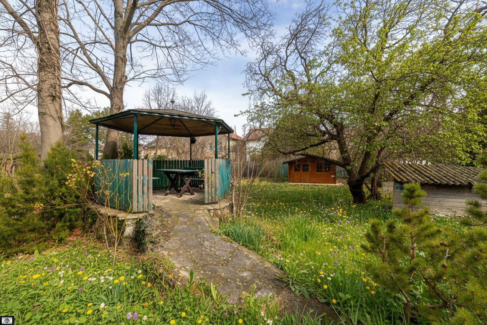 Idyllischer Gartenbereich mit einem hölzernen Pavillon, einem Gartenhaus und üppiger Vegetation.