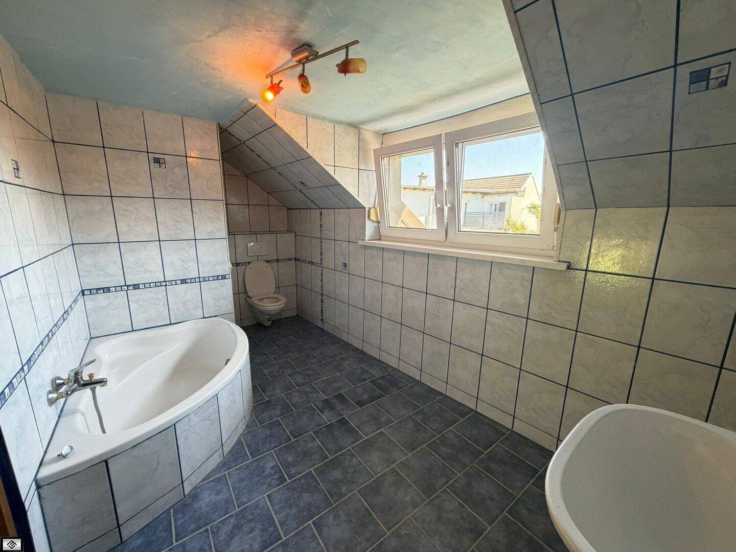 Badezimmer im Dachgeschoss mit blauen Fliesen, Eckbadewanne, Toilette und Fenster.