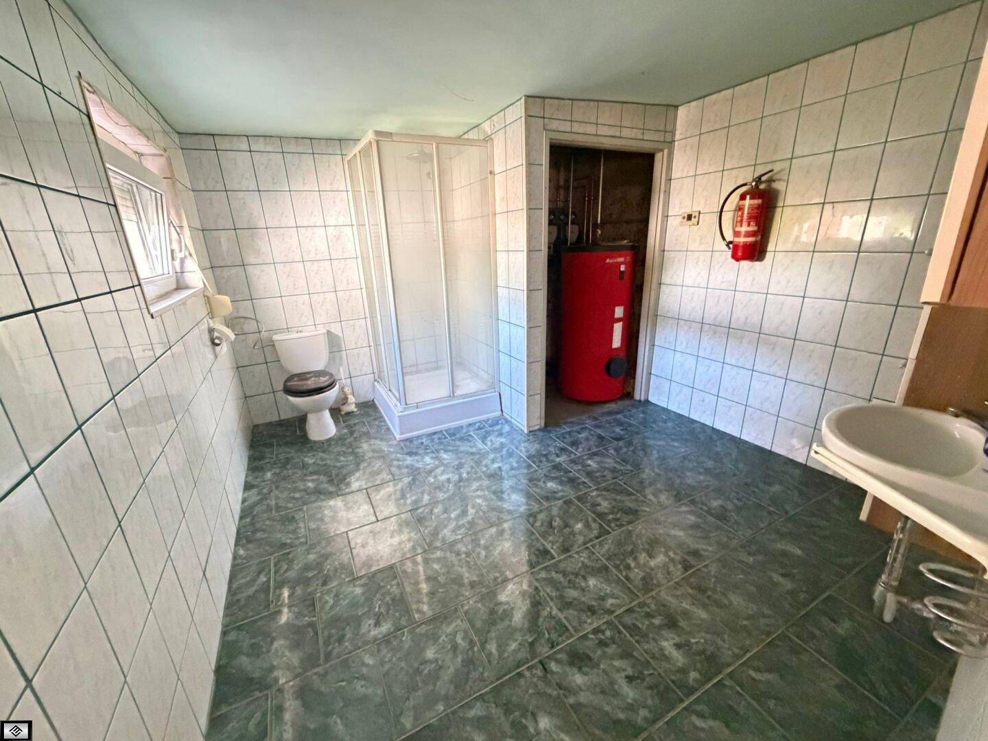 Gefliestes Badezimmer mit Dusche, Toilette, Waschbecken und einem Warmwasserboiler.