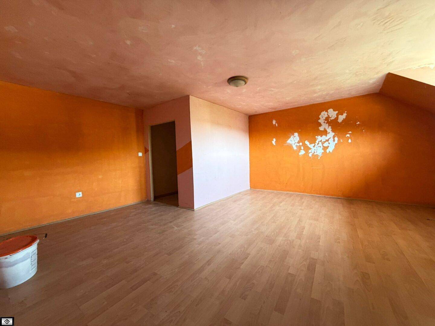 Leeres Zimmer mit orangefarbenen Wänden, Laminatboden und Dachschräge, renovierungsbedürftig.